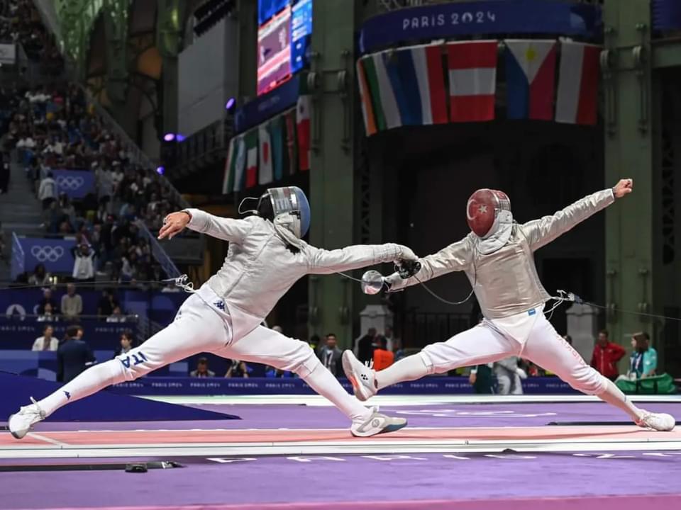 Two fencers, one wearing an ITA (Italy) uniform, lunge at each other during a bout at the Grand Palais, with national flags and the Olympic rings visible in the crowd behind them